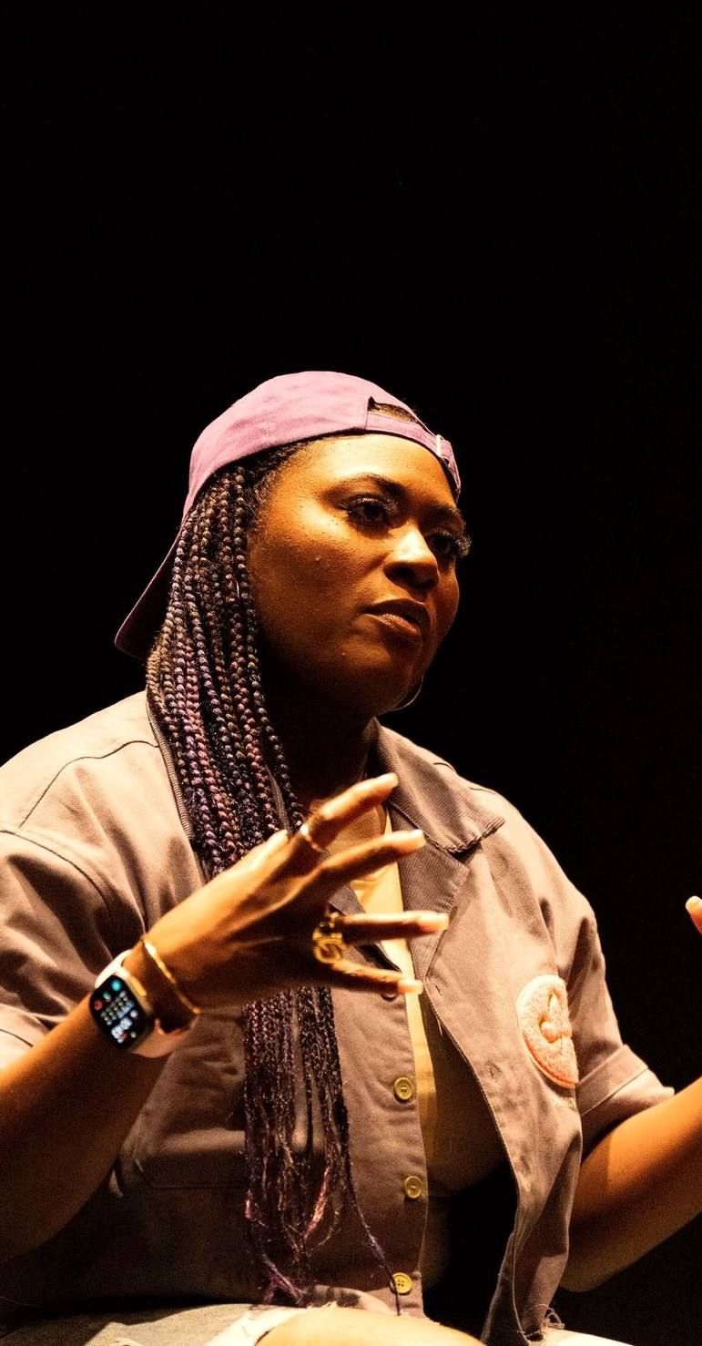 Danielle Brooks on the Governor's School stage, seated in a pool of light, mid-sentence.